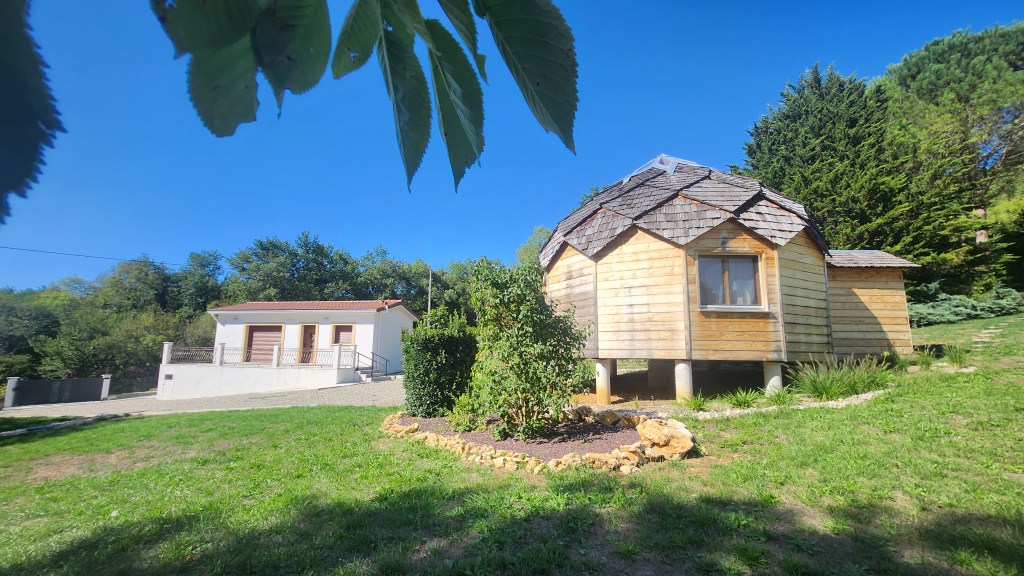 Vue d'une maison en bois avec un toit en ardoise, positionnée sur un terrain verdoyant, à côté d'une autre maison moderne.