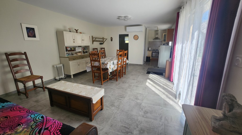 Intérieur d'un salon et d'une cuisine, avec des chaises en bois, une table, un meuble de rangement et un sol carrelé. Des fenêtres laissent passer la lumière.