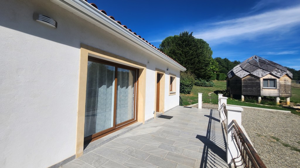 Vue d'une maison moderne avec un grand patio, des fenêtres en bois, et un abri en bois à proximité, sous un ciel bleu dégagé.
